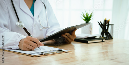 A professional female doctor, wearing a lab coat and stethoscope, sits at her desk with a tablet and clipboard, looking directly at the camera.
