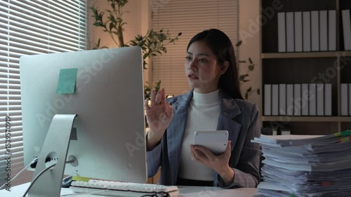 Dedicated young Asian businesswoman working late, analyzing complex project data on tablet and computer while managing extensive paperwork at dimly lit office desk