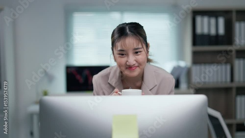 Office worker enjoying coffee break while working on computer, displaying cheerful expression at modern workplace with bright, professional interior