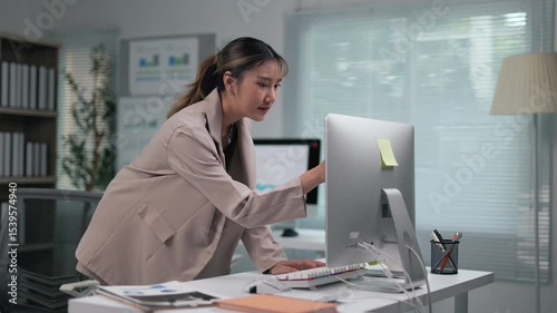 Young Asian businesswoman standing at standing desk, working diligently while typing on keyboard and analyzing data in modern corporate office environment