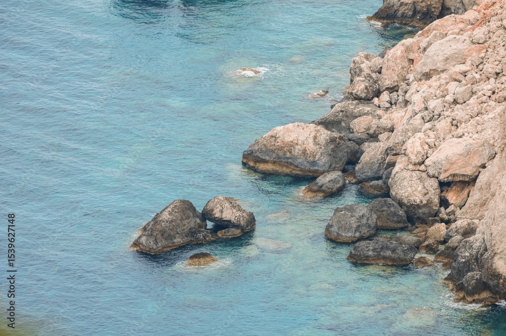 Fototapeta premium Adventurers discover rocky formations along a stunning shoreline, where waves gently lap against the boulders. Sunlight sparkles on clear water, inviting relaxation and exploration