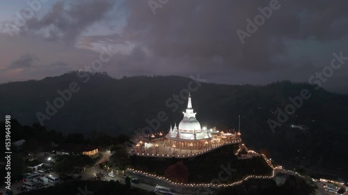 Hill top religious stupa decorativly lit up during blue hour on a mountain top in Kumbalwela, Ella, Sri Lanka. Drone dynamic lift aerial. photo