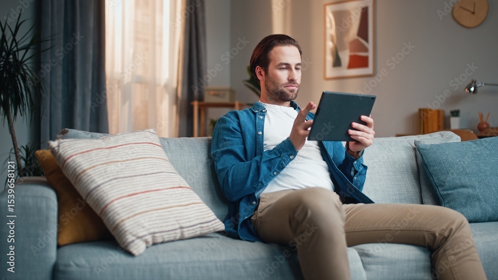Obraz premium Thoughtful man sitting on sofa and using tablet. Serious man putting legs on couch. Concentrated man using device and work at home. Male holding tablet and working distantly.