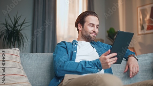 Busy charming man reading ebook using tablet online. Young beautiful male sitting on sofa in living room and reading interesting book smiling thinking. Stylish smart guy develops. Reading concept