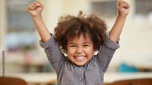 Young girl with curly hair is celebrating joyfully, raising her arms in excitement, showcasing a bright smile in a cheerful indoor environment, embodying happiness and youthful energy