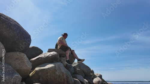 A man is peacefully relaxing on rocks that are next to the water on a beautiful day
