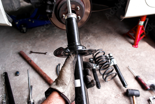  Car Shock Absorber or Strut, A mechanic hand holds a new black shock absorber or strut for replacement, surrounded by car parts and tools in a auto repair garage.