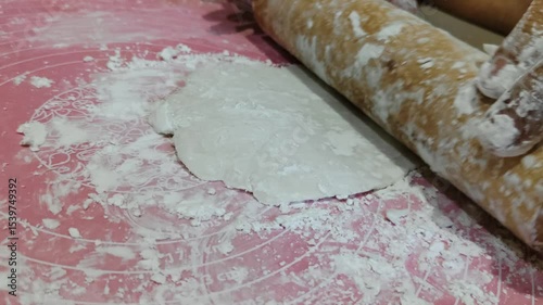 Hands actively rolling dough on a pink surface during food preparation. A workbench with cooking utensils indicates a domestic culinary activity, evoking a homely and creative atmosphere.
