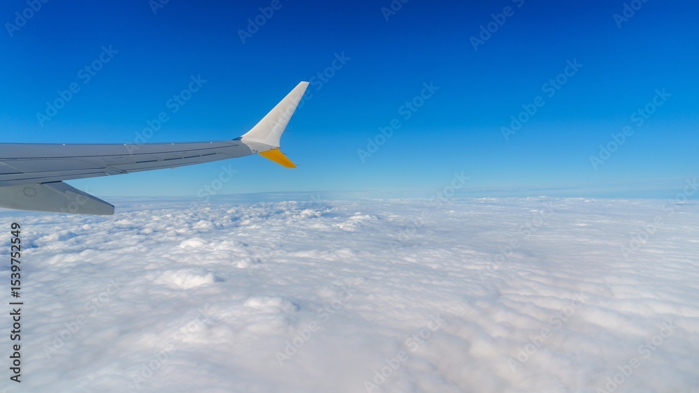 Fototapeta premium Stunning aerial view from an airplane window, featuring the aircraft s wing gliding above a sea of fluffy white clouds set against a bright blue sky. Perfect for travel and exploration.