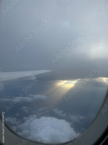 View from airplane window showing wing above clouds and sun rays breaking through dark sky. Concept of flight, sky travel, weather, and atmospheric beauty