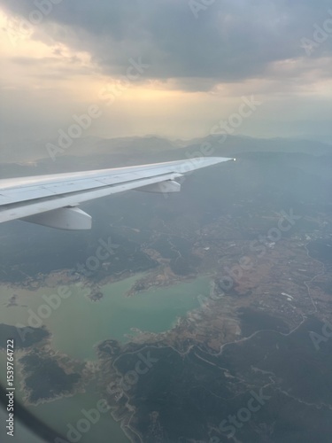 View from airplane window showing aircraft wing, mountainous landscape, and water reservoir below under cloudy sky. Concept of air travel, landscape observation, and tourism