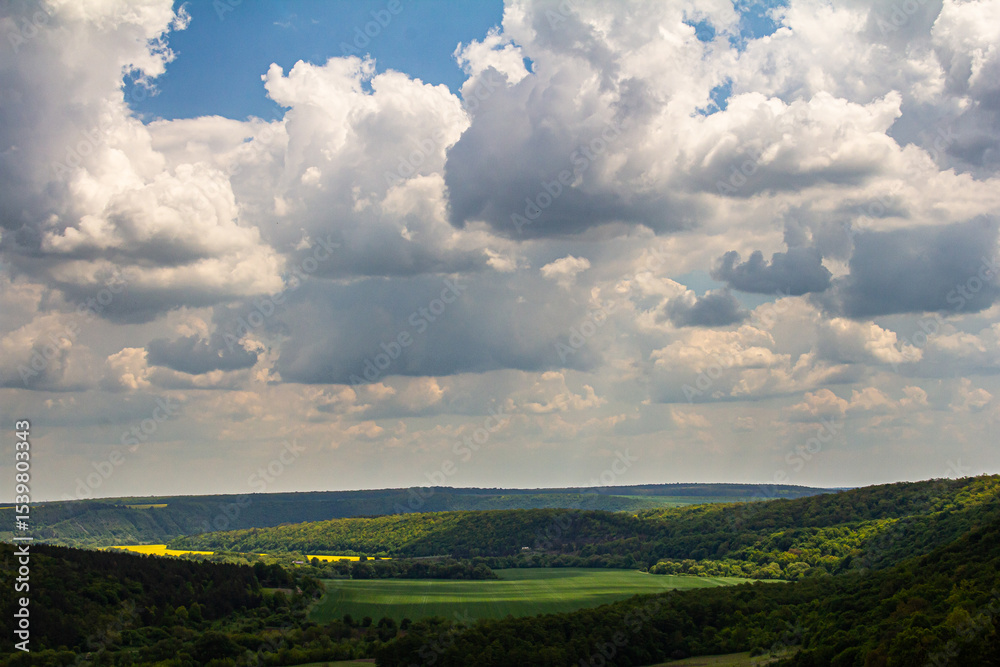 Fototapeta premium Scenic view of rolling hills and dramatic clouds over a lush green valley under a bright blue sky during midday