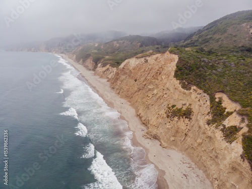Aerial view of a rugged coastline  of Point Reyes National Seashore, Bolinas, California, USA. Waves crash on the sandy beach below steep cliffs, with fog blanketing the hills in the background.