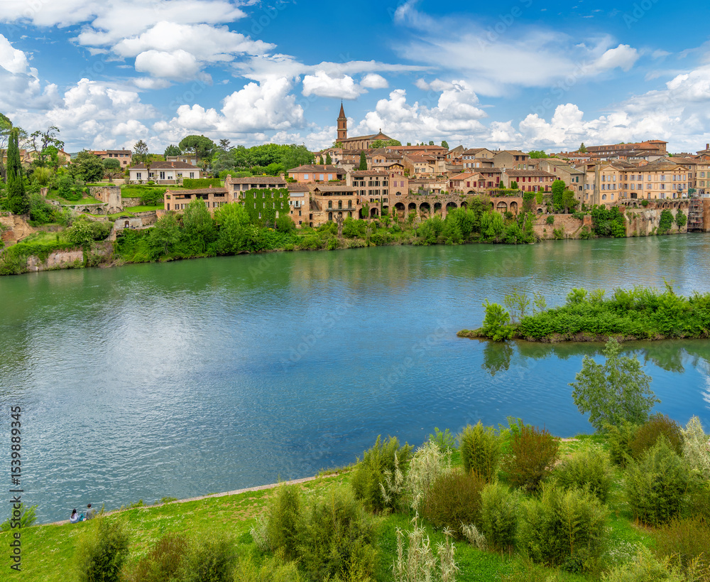 Fototapeta premium Panoramic view of the houses, historic architecture, and church of Albi, France, from the green Tarn River with a young couple in love having a picnic on the riverbank.