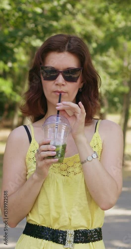 Fashionable woman stands in city park and sips refreshing mojito. Young female in bright yellow summer dress and sunglasses enjoys beauty of park. Vertical shot