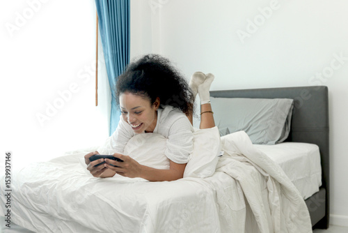 Beautiful curly hair woman in white bathrobe using smartphone after waking up in morning. African female checking mobile phone while lying down on comfortable bed at home. Happy girl surfing internet.