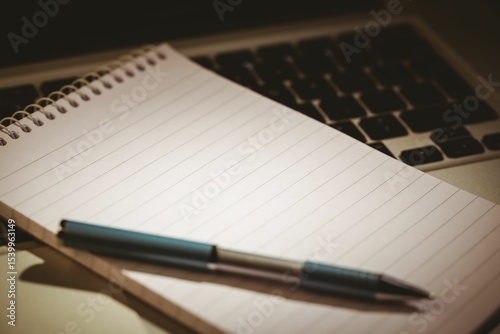 Flat design notebook with ballpoint pen is resting on laptop keyboard over desk with warm lighting