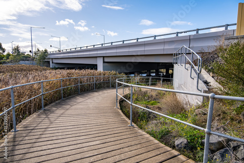 Behang A boardwalk curves through native wetland vegetation and passes beneath an overpass in suburban Australia