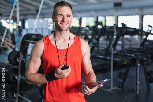 Mid adult trainer wearing athletic top holding stopwatch tablet in gym near weight benches, mirrors