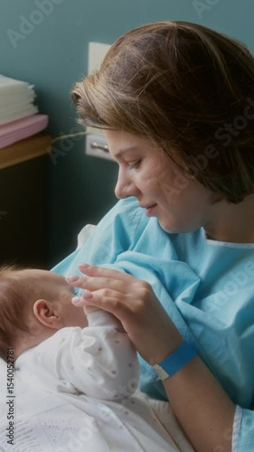 Happy Caucasian woman sitting on bed in maternity hospital room, touching small hand of her newborn baby
