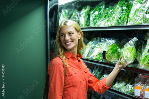 Woman wearing orange shirt carrying shoulder bag selecting lettuce in grocery store produce aisle
