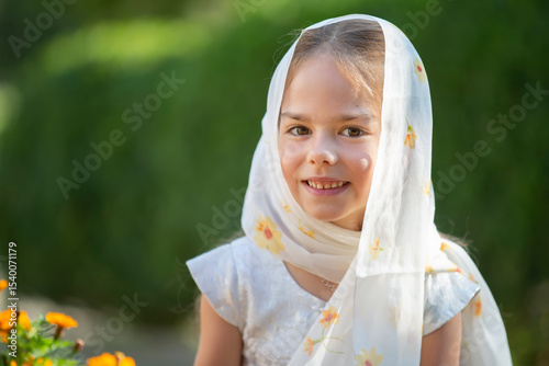 Young girl wearing a scarf smiles in a garden during daylight, surrounded by blooming flowers