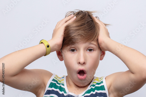 Young boy shows surprise and shock with hands on head in casual summer attire against a light background