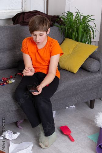 Boy in orange shirt sits on couch using smartphone in a messy living room