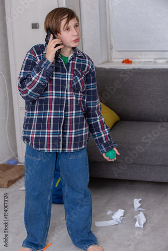 Boy in plaid shirt talking on phone while standing in messy living room with crumpled paper
