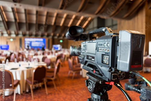 A professional video camera on a tripod set up in a spacious conference hall, facing round tables and a stage, ready to capture a business seminar or corporate event.