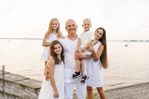 Happy family of five is posing for photo on the beach. Father is holding a baby, children are wearing white dresses