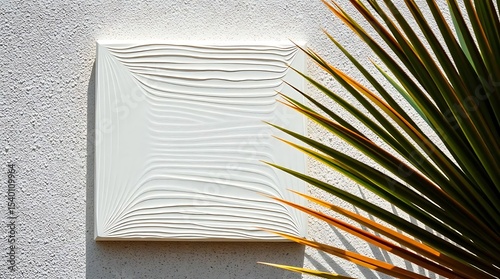 A white textured square art panel hangs on a textured wall with a palm leaf partially obscuring it