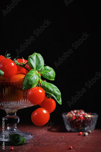 Artistic food still life of cherry tomatoes and basil leaves in vintage glass bowl on moody dark red surface