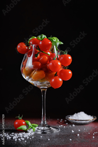 Red cherry tomatoes with basil in a glass, surrounded by salt on dark background, gourmet still life concept for clean eating and healthy meals