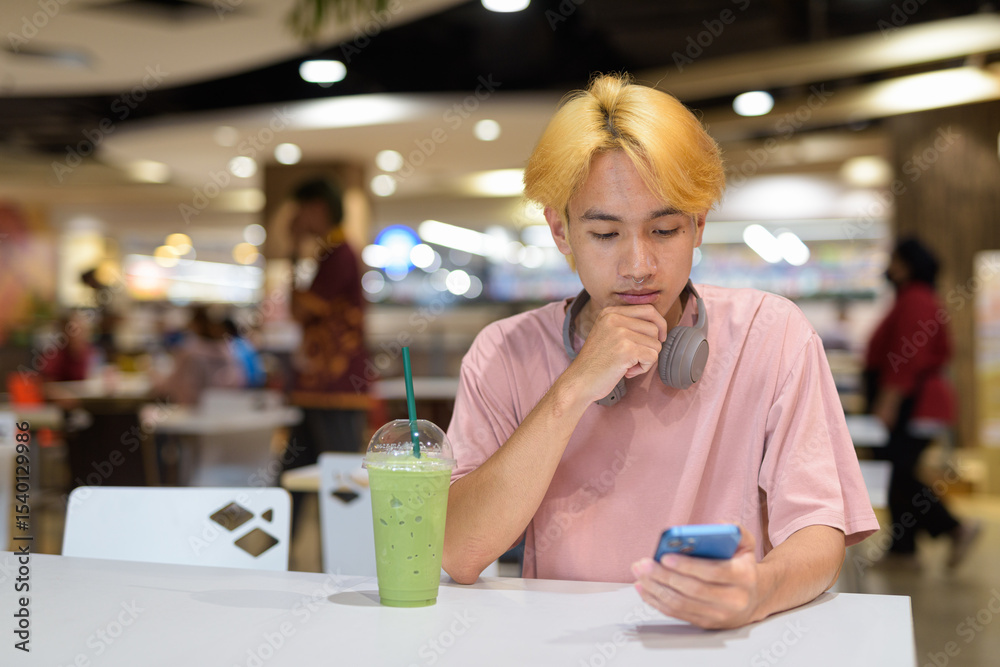 Fototapeta premium Thai gay teen man with headphones using phone and drinking iced latte green tea in modern cafe