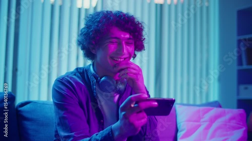 Mental Health, Digital Connection, Emotional Release. Young Man Sits on Sofa at Night, Laughing at Phone Screen. Moment of Joy, Relaxation, and Connection in Digital Daily Life