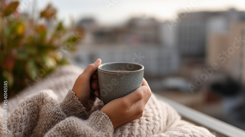 Gemütlicher Morgenmoment auf dem Balkon – warme Hände halten eine blaue Tasse Kaffee oder Tee in kuscheliger Kleidung mit Stadtblick und Pflanzen im Hintergrund