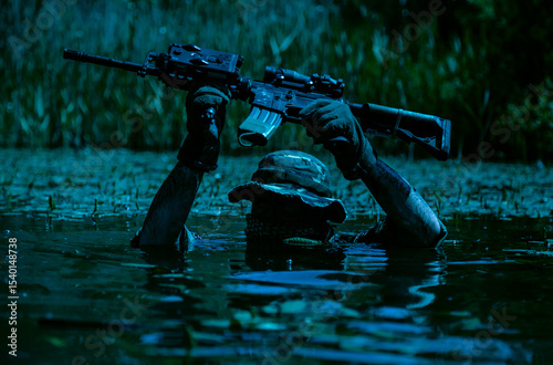 Canvas-taulu Soldier in camouflage gear and hat holding a rifle above water in a wetland area during nighttime