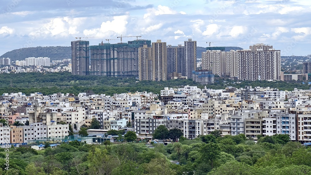 Fototapeta premium Dense urban landscape with a mix of low-rise and high-rise residential buildings set against a backdrop of green cover and hills under a cloudy sky.