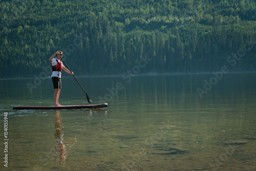Man wearing vest and holding paddle paddling on paddleboard in shallow lake by forest, copy space
