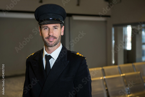 Mature male pilot standing near seats in airport lounge wearing peaked cap with striped epaulets