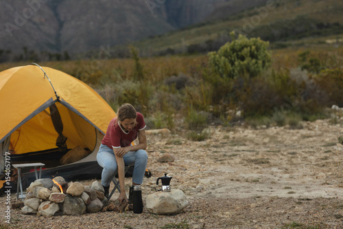 Woman camper leaning forward beside stone fire pit and yellow tent at campsite reaching for kettle