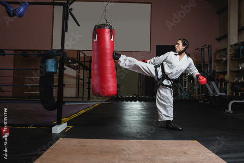 Female martial artist in white gi delivering kick at gym with red punching bag, copy space
