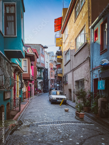 Narrow cobblestone Istanbul Balat street winding past colorful homes and parked vintage car, lined with balconies, plants, and urban charm in a quiet old district.