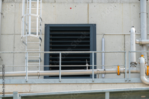 Modern factory view with metal piping, vent grating, ladder for service workers to climb up, on a concrete wall.