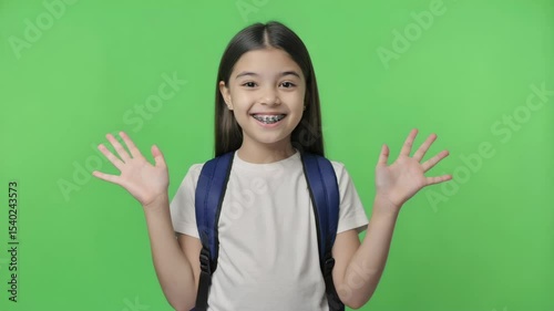 Smiling young girl with long dark hair, wearing a backpack, raises her hands in excitement against a green screen background.