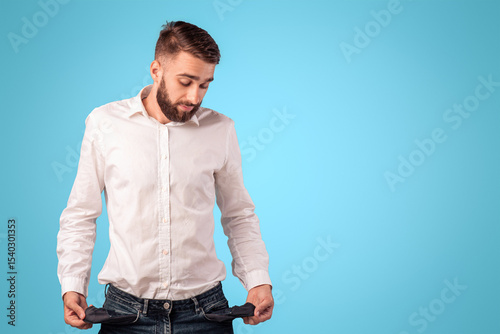 Young bearded man showing empty pockets on blue studio background. Millennial guy losing money, facing bankruptcy or unemployment during financial crisis. Economic depression concept