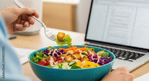A person eating a healthy and nutritious salad from a bowl while working on a laptop at a desk a concept of a modern office lunch wellness and a balanced diet lifestyle with fresh vegetables