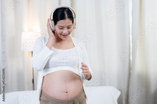 Asian pregnant woman smiling wearing headphones standing happily enjoying music with hand resting on belly expressing excitement bonding emotional connection with unborn baby at cozy home