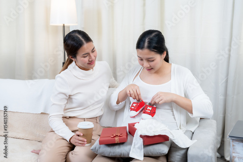 Asian pregnant woman happily opening baby gift box from friend sitting together on sofa expressing joy excitement appreciation celebrating baby’s arrival wearing casual white sweater brown pants